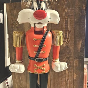 May include: A Sylvester the Cat nutcracker figure dressed in a red military-style uniform with gold fringe epaulets, a black belt, and a red nose. The figure features a tall hat topped with a yellow Tweety Bird head and white feathers.