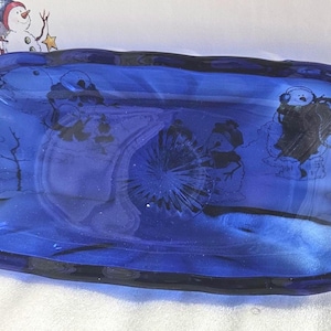 Puede incluir: Una fuente de vidrio azul oscuro con un borde ondulado. El plato tiene un diseño de escena invernal grabado en el vidrio. El diseño incluye muñecos de nieve y árboles. El plato mide aproximadamente 30 cm de largo y 15 cm de ancho.