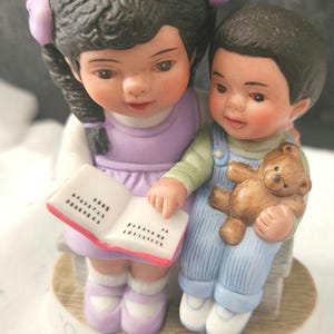 May include: A porcelain figurine of two children, a girl with purple pigtails and a boy, sitting on a bench. The girl is reading a book and the boy is holding a teddy bear. The base of the figurine reads "Our special story".