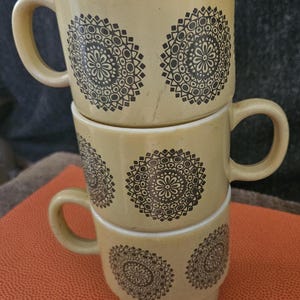 Könnte beinhalten: Drei übereinander gestapelte Keramikbecher in Braun- und Gelbtönen. Jeder Becher hat ein schwarzes Mandala-Design.