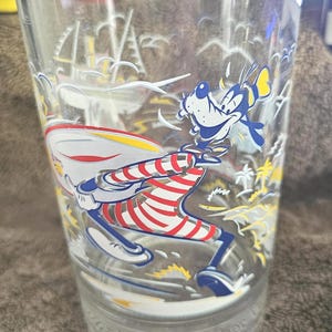 Puede incluir: Vaso de vidrio transparente con una imagen de dibujos animados de Goofy con un atuendo rojo, blanco y azul, con un avión a juego. El vaso tiene un diseño de isla tropical y las palabras "World Remember" en la base.