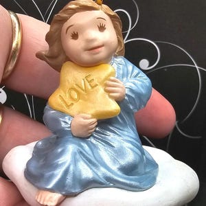 Puede incluir: Figura de un ángel niño con una túnica azul, arrodillado sobre una nube blanca. El ángel sostiene un objeto amarillo en forma de estrella con la palabra "LOVE" escrita. La figura tiene cabello castaño y un halo dorado.
