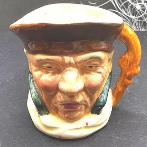 May include: A vintage ceramic mug shaped like a man's face, featuring a brown hat, a tan face with rosy cheeks, and a white collar. The handle is a light orange color. The mug is a unique collectible.