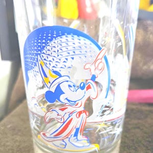 May include: Clear glass with a colorful graphic of Mickey Mouse in a sorcerer's outfit, with a blue and white background. The glass has the words "Remember The Magic" at the bottom.