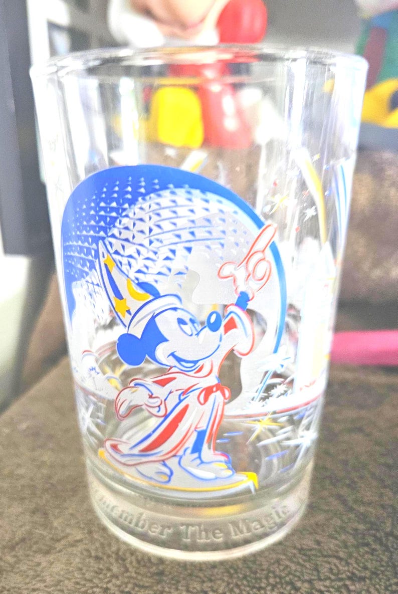 May include: Clear glass with a colorful graphic of Mickey Mouse in a sorcerer's outfit, with a blue and white background. The glass has the words "Remember The Magic" at the bottom.