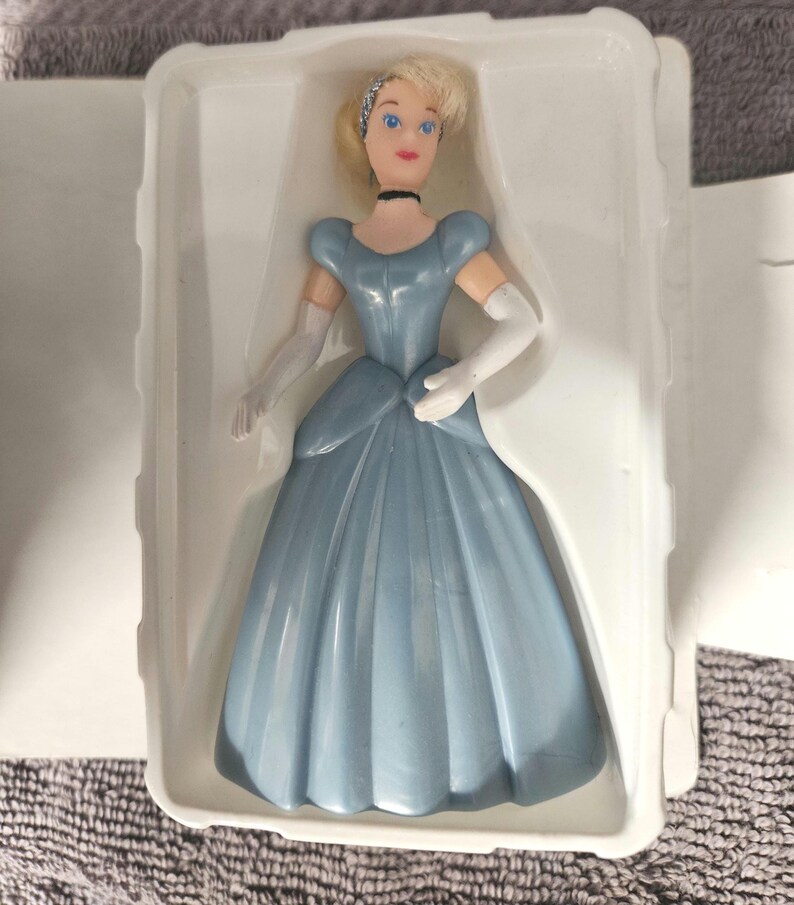May include: A plastic Cinderella doll in a blue ball gown with white gloves and a tiara. The doll is in a white plastic package.
