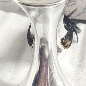 May include: A silver-colored, decorative wine bottle stopper with a flared top and a base. The stopper features black and gold accents, with a feather-like design. The background shows a winter scene with snowmen.