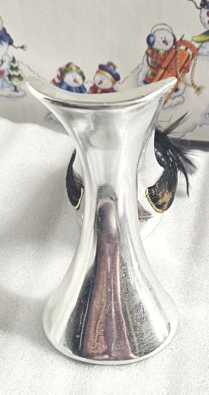 May include: A silver-colored, decorative wine bottle stopper with a flared top and a base. The stopper features black and gold accents, with a feather-like design. The background shows a winter scene with snowmen.