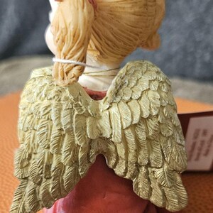 以下が含まれることがあります： 金色の羽根を持つ、赤いドレスを着た金髪の天使の小さな置物。天使は頭を下げて座り、羽根は後ろに広げられている。