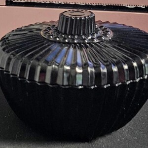 May include: A black, vintage-style trinket box with a ribbed, oval-shaped body and a decorative, ridged lid. The lid has a small, circular handle. The box is made of a glossy material, reflecting light. The overall design is elegant and classic.