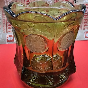 Pode incluir: Um vaso de vidro verde com uma borda festonada e uma superfície texturizada. O vaso tem um desenho de medalhões circulares com um padrão de estrela.