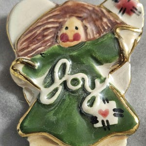 May include: A ceramic angel figurine with a green dress and brown hair. The word "joy" is written across the front. The angel has a gold trim and a small heart on the front.