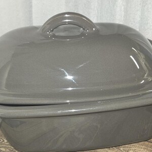 May include: A gray ceramic casserole dish with a lid and a handle. The dish is oval-shaped and appears to be made of a glossy ceramic material. The lid has a loop handle for easy lifting.