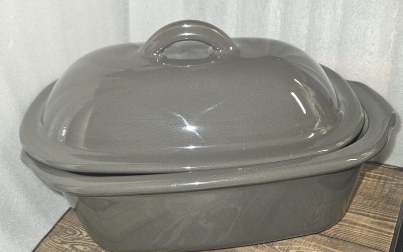 May include: A gray ceramic casserole dish with a lid and a handle. The dish is oval-shaped and appears to be made of a glossy ceramic material. The lid has a loop handle for easy lifting.