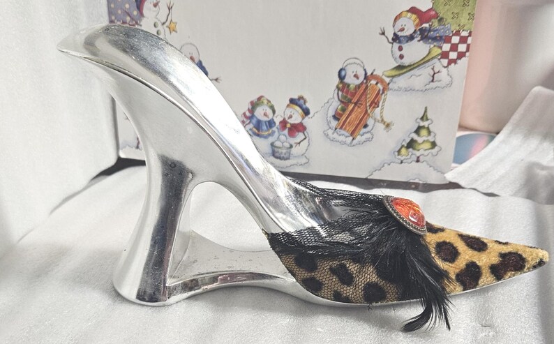 May include: A decorative silver high-heeled shoe with a leopard print toe and black feather accents. The shoe features a decorative jewel and black lace detailing. The heel is a unique, open design.
