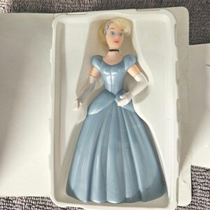May include: A plastic Cinderella doll in a blue dress with white gloves and a tiara. The doll is packaged in a white plastic clamshell.