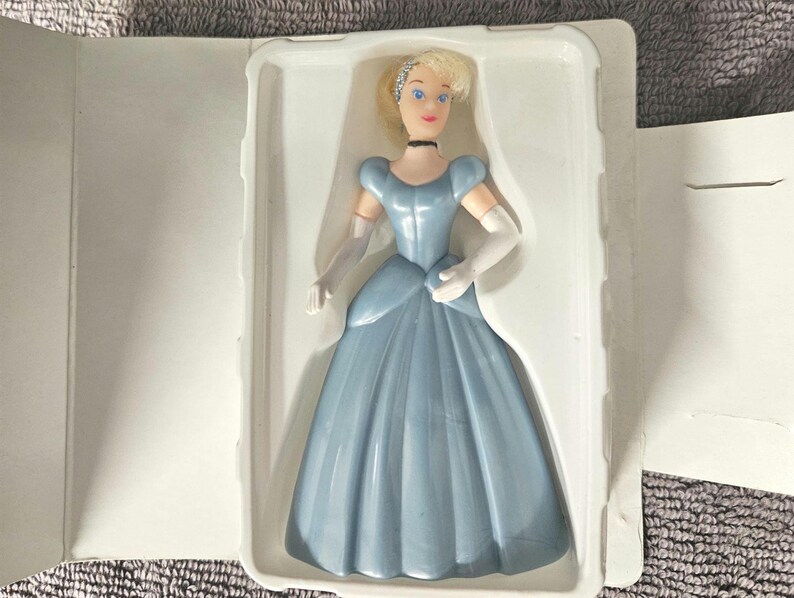 May include: A plastic Cinderella doll in a blue dress with white gloves and a tiara. The doll is packaged in a white plastic clamshell.