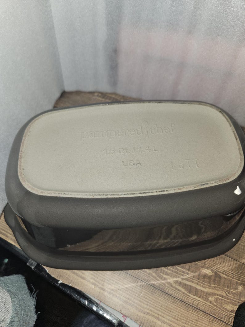 May include: A dark gray rectangular Pampered Chef baking dish. The bottom of the dish is light gray with the words "pampered chef", "1.5 QT/1.4 L", and "USA" printed on it. The dish has a capacity of 1.5 quarts or 1.4 liters.