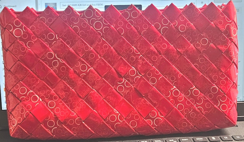 K&ouml;nnte beinhalten: Eine rote gewebte Clutch mit Rei&szlig;verschluss. Die Clutch besteht aus einem gl&auml;nzenden Material mit einem Muster aus wei&szlig;en Kreisen. Das Gewebe ist diagonal und erzeugt eine strukturierte Oberfl&auml;che. Der obere Rand hat ein Zickzack-Design.