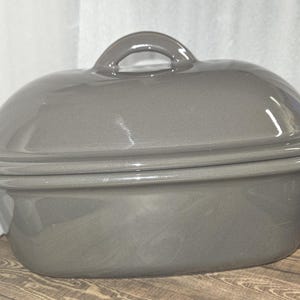 May include: A gray ceramic oval casserole dish with a matching lid. The lid has a loop handle. The dish is sitting on a wooden surface.