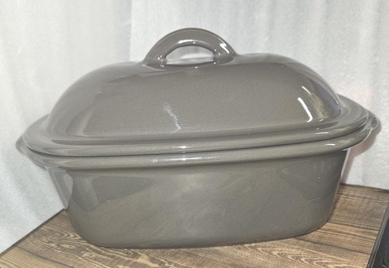 May include: A gray ceramic oval casserole dish with a matching lid. The lid has a loop handle. The dish is sitting on a wooden surface.