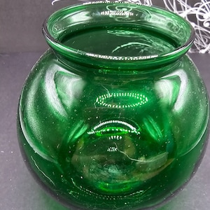 May include: A round, green glass jar with a wide opening. The jar is made of thick glass and has a smooth, shiny surface.