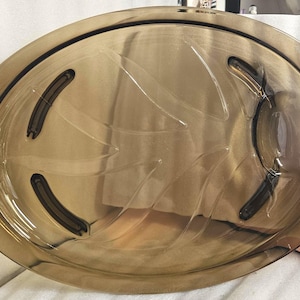 Puede incluir: Una fuente ovalada de color ámbar. El diseño transparente presenta detalles curvos y cuatro hendiduras curvas de color marrón oscuro. Los bordes están ligeramente elevados con detalles decorativos.