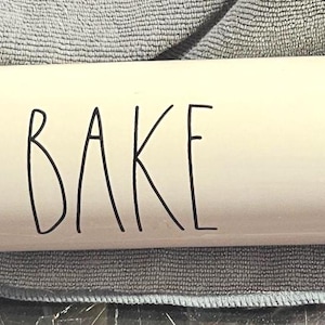 May include: A cream-colored rolling pin with the word "BAKE" in black lettering. The handles are made of light-colored wood. This kitchen tool is designed for baking.