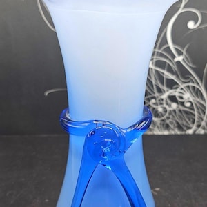May include: A light blue glass vase with a flared, ruffled rim. The vase has a cylindrical body with a darker blue, decorative band and accents. The design includes a swirl detail on the band and flowing lines.