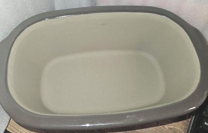 May include: A gray ceramic baking dish with a rectangular interior and rounded corners. The dish has a light gray interior and a darker gray rim and handles. This kitchenware is suitable for baking and serving.