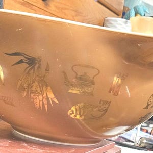 Puede incluir: Un gran cuenco para mezclar de color marrón claro con una boquilla. El cuenco está decorado con ilustraciones doradas de un gallo, plumas, una tetera y otros diseños temáticos de cocina. El cuenco está sobre una superficie roja.