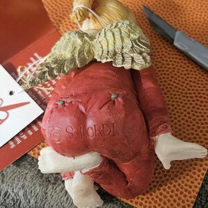 以下が含まれることがあります： 赤いセラミック製の天使の置物で、金色の羽根が付いています。赤い服を着ており、白い縁取りが施されています。置物は、足を曲げてお尻に座っています。置物の底には「S LORDI」と署名されています。
