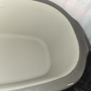May include: A close-up view of a ceramic baking dish. The interior is a light cream color, and the rim is a darker gray. The dish has a rectangular shape with rounded corners. The surface appears smooth.