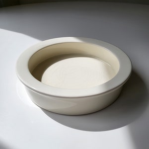 May include: A white ceramic dish with a rounded rim and a smooth, slightly textured surface. The dish is empty and appears to be made for holding small items.