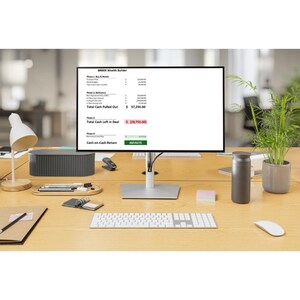 May include: A computer monitor displaying financial data sits on a wooden desk. A white keyboard, mouse, and a small desk lamp are in the foreground. Other items include a gray speaker, a stapler, a pen, a notebook, a thermos, and a potted plant.