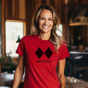 May include: A red t-shirt with two black diamond shapes and the word "DIFFICULT" in a black font. The short-sleeved shirt is made of a soft material. The design is centered on the chest.