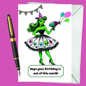 Puede incluir: Tarjeta de felicitación de cumpleaños con una mujer alienígena verde con un vestido negro y una falda blanca con diseños de alienígenas. La tarjeta incluye el texto "Hope your birthday is out of this world!" y va acompañada de un bolígrafo y un sobre.