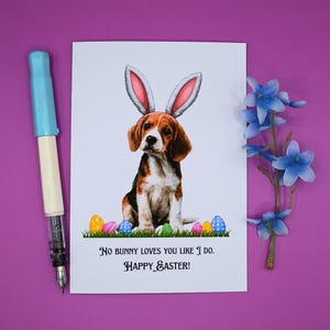 May include: An Easter greeting card featuring a beagle wearing bunny ears, surrounded by colorful Easter eggs on grass. The card includes the text "No bunny loves you like I do. Happy Easter!" A blue pen and blue flowers are also in the image.