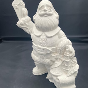 May include: A white ceramic Santa Claus figurine holding a letter and a sack. The Santa figure has a long white beard, a belt, and a fur-trimmed coat. The sack has a bell and a scroll with text.