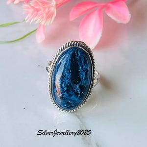 May include: Oval-shaped silver ring with a large, dark blue gemstone. The gemstone has a marbled appearance with lighter blue and white swirls. The ring is set against a white surface with pink flowers in the background. The text "SilverJewellery2025" is visible.