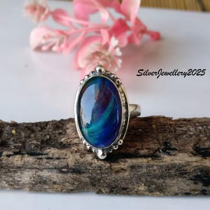 May include: A silver ring with an oval-shaped, iridescent blue and green stone. The stone is set in a decorative silver bezel. The ring is displayed on a piece of wood with pink flowers in the background. The text "SilverJewellery2025" is visible.