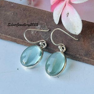 Puede incluir: Un par de pendientes de plata con piedras preciosas ovaladas de color azul claro. Los pendientes tienen un diseño sencillo con cierre de gancho. Las piedras preciosas están engastadas en un bisel de plata. Los pendientes se exhiben sobre una superficie de madera.