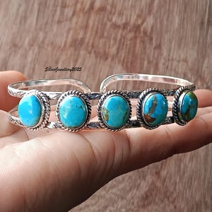 May include: A silver cuff bracelet featuring five oval turquoise stones set in twisted rope borders. The metal band has an etched pattern and the text SilverJewellery2025 is visible. The bracelet has an open back design with a polished finish.