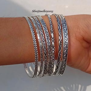 May include: Set of seven silver-toned bangles with intricate floral and geometric designs. The bangles are stacked on a person's wrist.