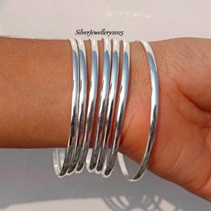 May include: A set of eight silver bangles, stacked on a person's wrist. The bangles are smooth and shiny, with a polished finish.
