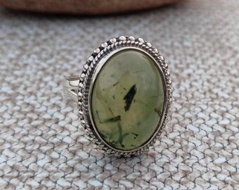 Könnte beinhalten: Ein silberner Ring mit einem ovalen grünen Edelstein, der in einer Perlenfassung sitzt. Der Stein hat eine durchscheinende grüne Farbe mit dunkelgrünen Einschlüssen.