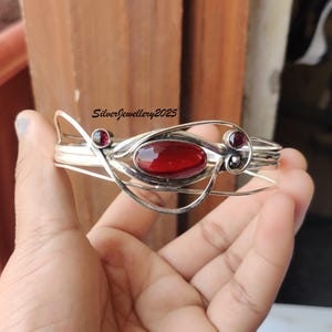 Pode incluir: Um bracelete de prata com uma pedra oval vermelha e duas pedras vermelhas menores. O bracelete é feito de fio de prata trançado e tem um design delicado e intrincado.