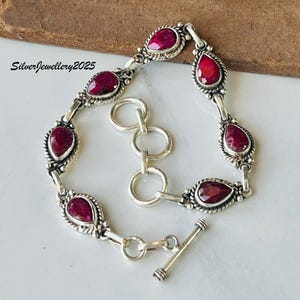 Könnte beinhalten: Silberarmband mit tropfenförmigen, tiefroten Edelsteinen. Das Armband hat ein verziertes Design im Vintage-Stil mit einem Knebelverschluss. Der Text "SilverJewellery2025" ist sichtbar.