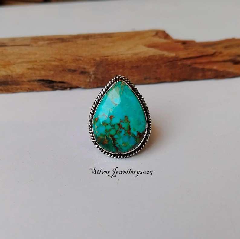 May include: A turquoise teardrop-shaped ring set in silver with a rope-like border. The ring is on a wooden surface.