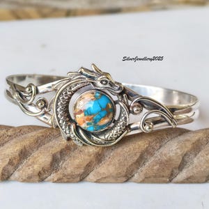 Könnte beinhalten: Silberarmband mit Drachenmotiv. Das Armband hat einen türkisfarbenen und kupferfarbenen Stein in der Mitte, der vom Körper des Drachen umrahmt wird. Der Drachenkopf befindet sich oben, und das Armband hat eine polierte Oberfläche. Der Text "SilverJewellery2025" ist sichtbar.